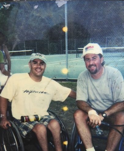 Two wheelchair tennis players sit together beside an outdoor tennis court, highlighting friendship, camaraderie, and the community built through adaptive tennis.